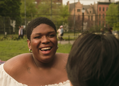 A non-binary trans woman laughing in a park.png